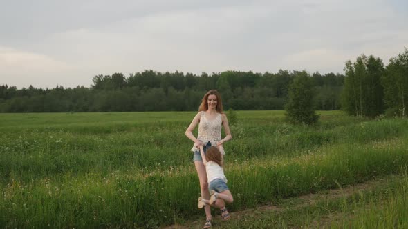 Young Mother Twists a Baby Girl on a Green Field in Summer