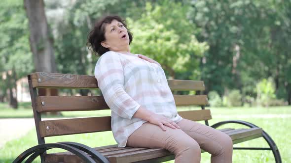 Ill Caucasian Senior Woman Having Heart Attack Sitting on Bench in Summer Park As People Passing By