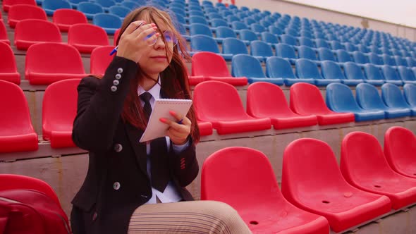 Young Woman in Glasses with Notepad Pen Sitting on Stadium Bleachers Alone