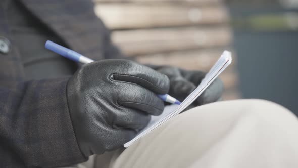 Close-up of Male Hand in Leather Glove Writing with Pen in Notebook. Unrecognizable Intelligence