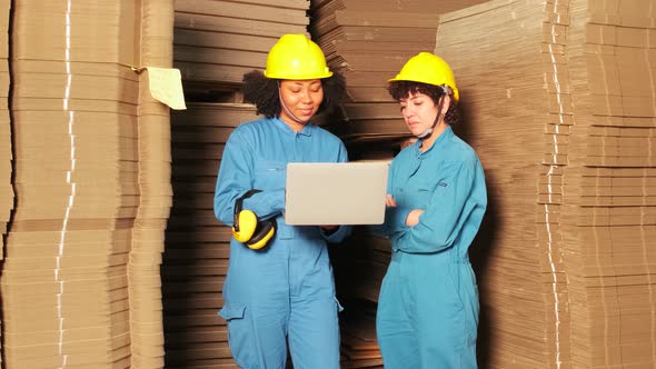 Two female workers in safety uniforms check pile of stock in cardboard factory.