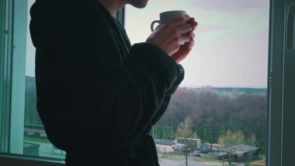 Man Drinks Coffee From Cup in Black Bathrobe at an Open Window in the Morning