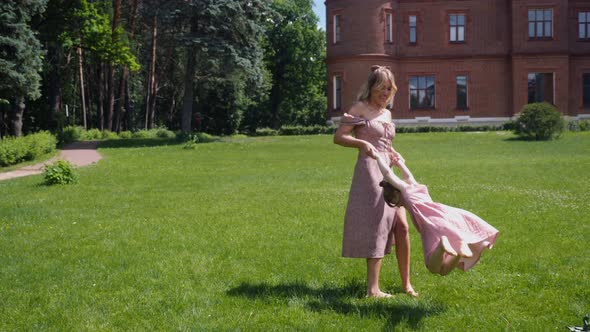 Woman and a Child in Pink Dresses Run Across a Green Field Against the Background of an Old Castle