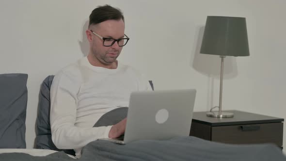 Casual Man Smiling at Camera While Using Laptop in Bed