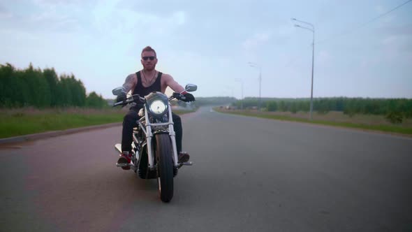 Stylish Biker with Tattoos Rides a Motorcycle on a Country Road at Sunset