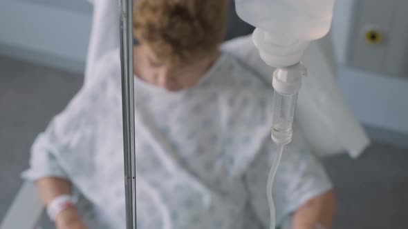 Close up saline solution drip, elderly lady resting on hospital chair blurred background