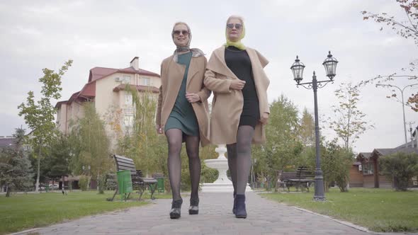 Two Elegant Mature Caucasian Female Friends in Beige Coats, Short Dresses and Sunglasses Walking in