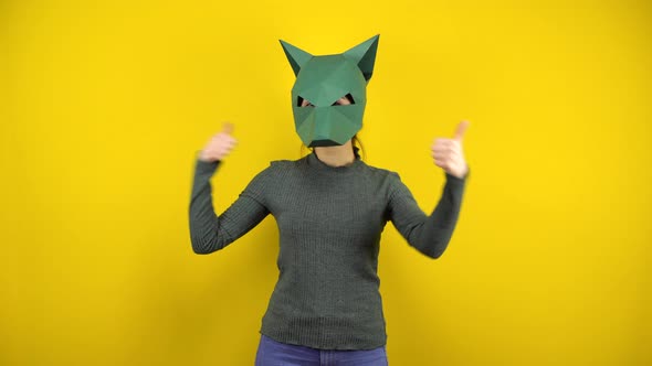 A Young Woman in a Cardboard Jackal Mask Shows Her Thumbs Up on a Yellow Background. Woman in Green