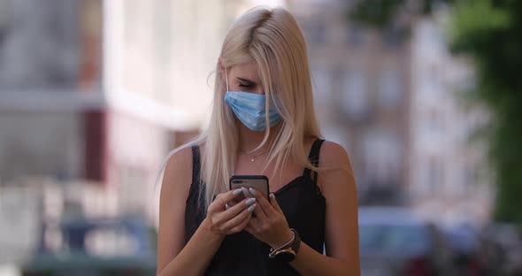 Young Blonde Woman in Protective Medical Mask Walks Down To the Street Uses Phone Texts Scrolls