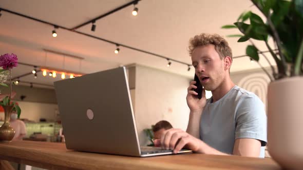 Young man working on laptop indoors. He talks to someone on the phone