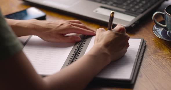 Woman's Hands Writing on Paper Notebook Page Sitting with a Cup of Coffee and Laptop in Cafe