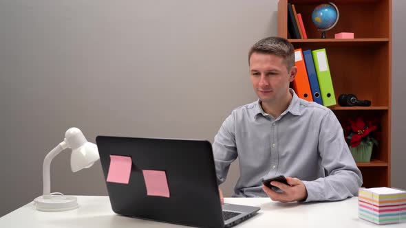 Young Guy Office Worker Manager is Typing Text in the Phone and in the Computer