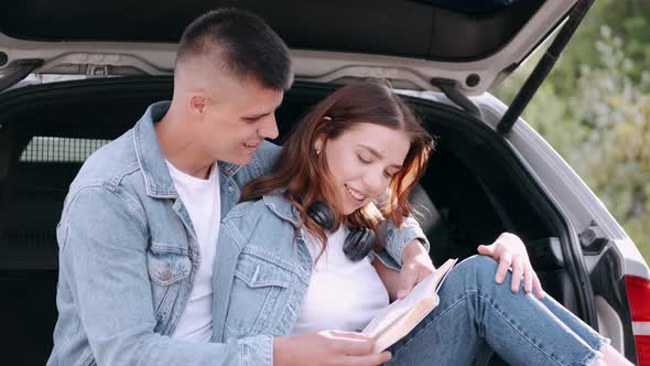 Couple Sitting in Car Trunk and Reading Book Together