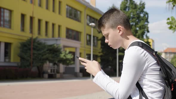 A Caucasian Teenage Boy Works on a Smartphone  Side View  a School in the Background