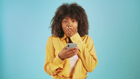 Afro American Female Looking at Mobile Phone Covering Mouth By Hand Being Surprised Rejoicing and