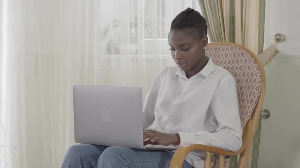 Attractive Afican American Woman Focused Working with Her Up To Day Laptop Sitting on Armchair