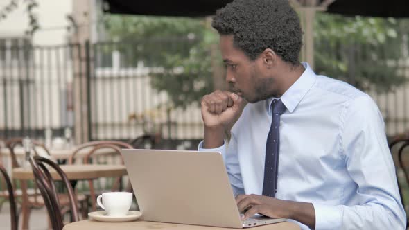 Coughing African Businessman Working on Laptop in Outdoor Cafe