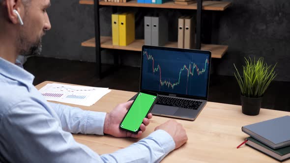 Man Stock Trader Looks Green Screen Smartphone Laptop with Stock Exchange Chart