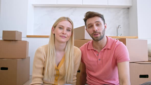 Couple in love lady and guy look straight and wave hands sitting near large brown cardboard boxes