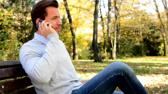 Young Man Sits on Bench in a Park and Talks To Somebody on the Phone