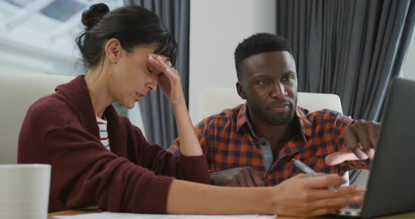 Diverse couple sitting at table talking and working with laptop