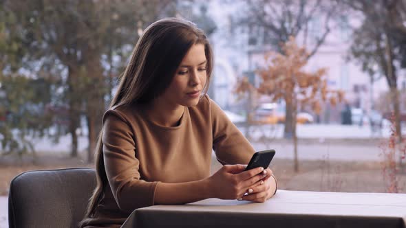 Young Woman Browsing on Smartphone Sitting Alone at Cafe Near Window