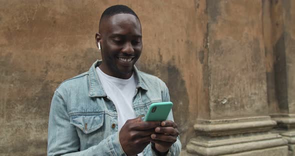 Close Up Shot of African American Guy in Jean Jacket with Earphones Using His Smartphone and