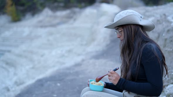 Girl in Hat and Glasses Sits in Stony Nature and Eats From Lunch Box