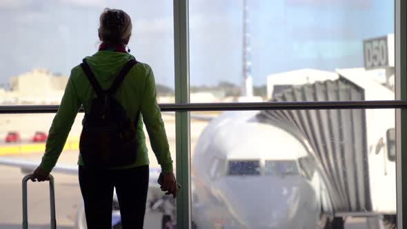 With blurry airplane at boarding gate, silhouette of woman walking over to glass and using her smart