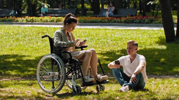 Woman in a Wheelchair and Her Friend Spending Time at the Summer Park and Sitting in Their Phones