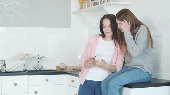 Two Excited Caucasian Women Surfing Internet on Smartphone. Middle Shot of Happy Lesbian Couple