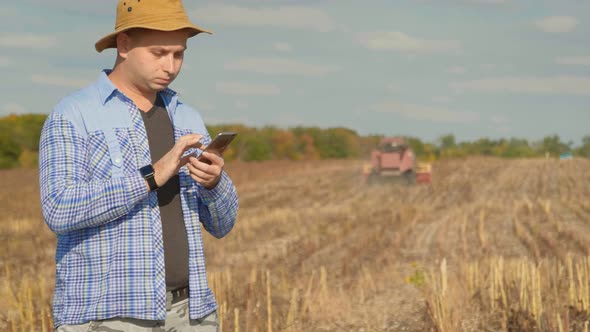 Handsome Farmer with Smartphone Standing in Field Sunflower with Combine Harvester in Background