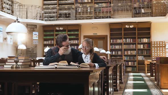 Man in Glasses Sitting at the Library Table and Listening How Read his Lovely Teen Granddaughter