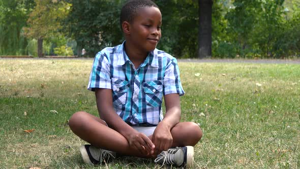 A Young  Black Boy Sits on Grass in a Park and Dances with a Smile