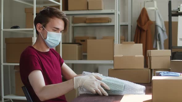 Man Employee of Warehouse Wearing Medical Mask and Gloves Packing Vase Into Wrapping Paper and
