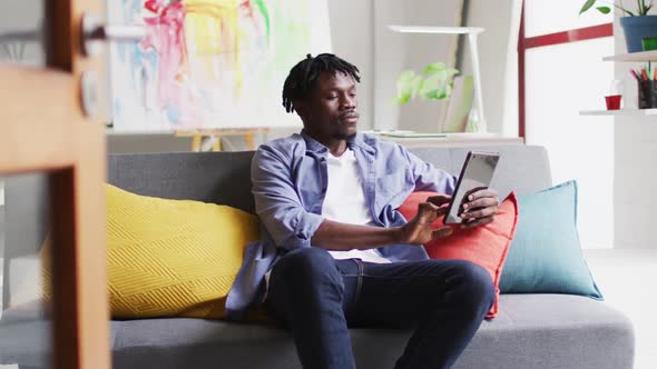 African american male artist using digital tablet while sitting on the couch at art studio