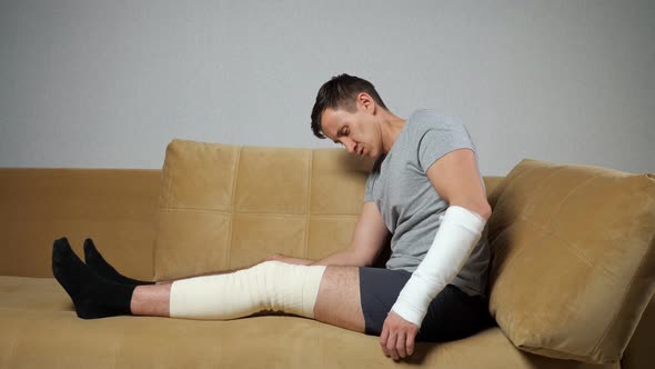 Man Sits on Sofa with Injured Knee and Broken Forearm