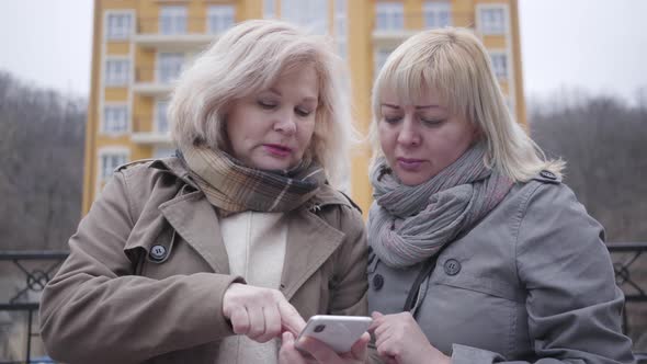 Close-up of Two Senior Caucasian Women Standing on City Street with Smartphone and Talking. Lost