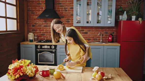 Smiling Little Girl is Cutting Lemon on Wooden Board Standing By Table in Kitchen at Home