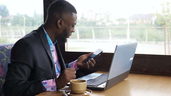 Black Businessman Works on Laptop with Smartphone in Cafe and Drinks Coffee.