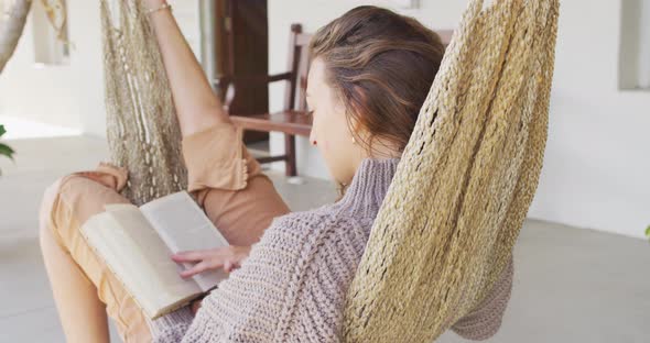 Smiling caucasian woman lying in hammock reading book on terrace