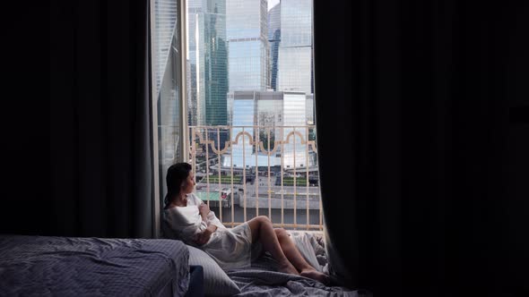 Woman in a Bathrobe Sits on the Floor of a Balcony Against the Backdrop of a Skyscraper During the