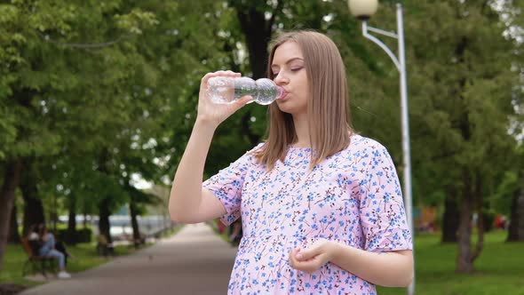 A Pregnant Woman in a Summer Dress with a Floral Print Stands in the Park and Drinks Pure Spring