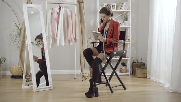 Wide Shot of Confident Young Brunette Fashion Designer Sitting in Atelier with Tablet and Talking on