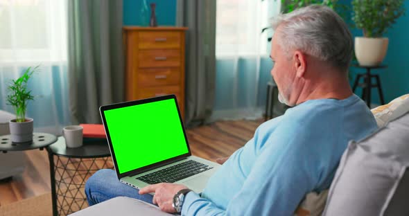 Man Uses Laptop with Green Mock Up Screen Sitting on a Couch at Home