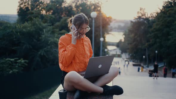 A Hardworking and Optimistic Girl Talks on the Phone Smiles Broadly While Typing on the Laptop