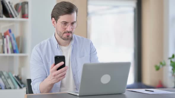 Man in Glasses with Laptop Using Smartphone in Office