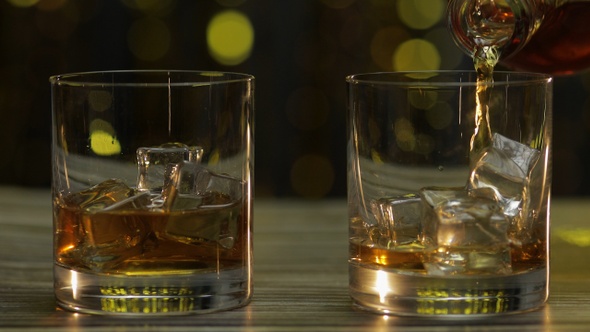 Pouring of Whiskey, Cognac or Brandy From Bottle Into Glasses with Ice Cubes. Shiny Background