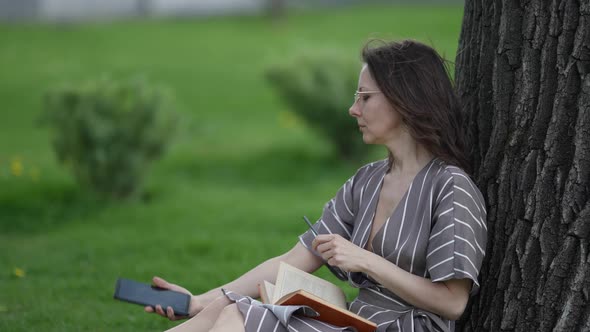 Pretty Woman is Reading Book and Taking Selfie By Camera of Smartphone Sitting Under Tree in Park