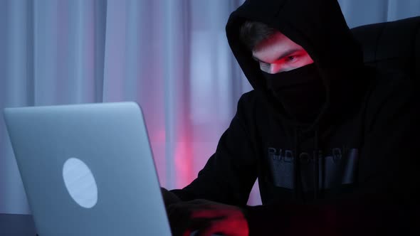 Hacker face in black mask and hood looking at laptop screen sitting in dark room